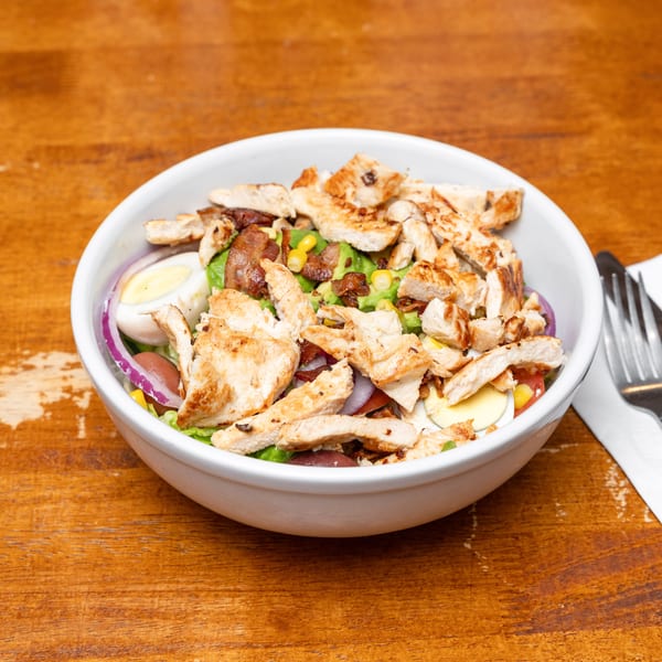 White bowl of grilled chicken salad with avocado, bacon, egg, and red onion on a wooden table.
