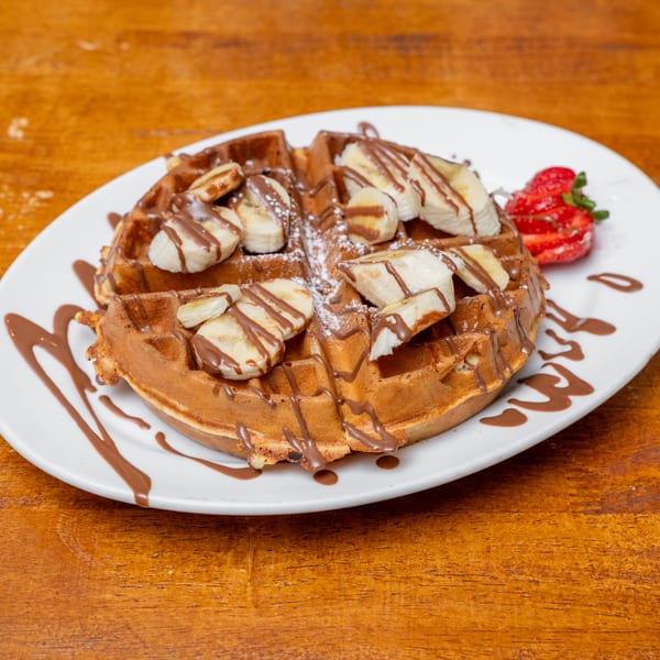 Golden waffle with banana slices, chocolate drizzle, powdered sugar, and strawberries on a wooden table.