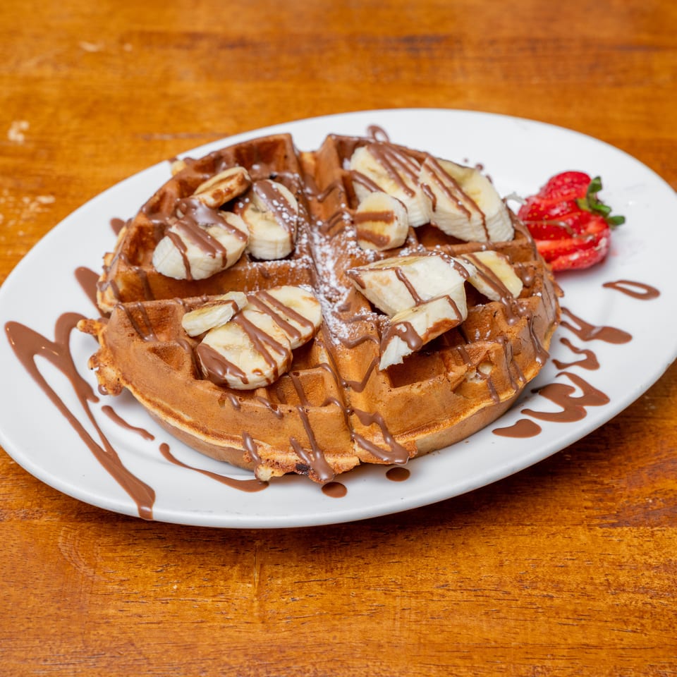 Golden waffle with banana slices, chocolate drizzle, powdered sugar, and strawberries on a wooden table.