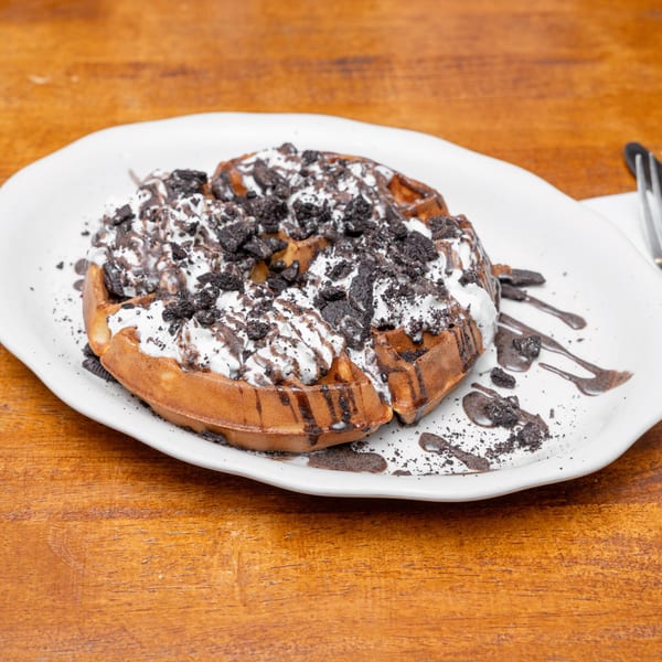 Waffle with whipped cream, chocolate drizzle, and crushed cookies on a white plate, on a wooden table.